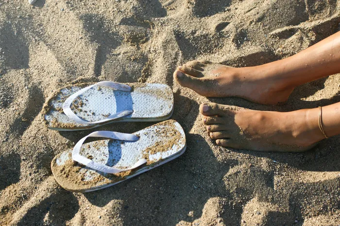A persons feet next to their white flip-flops resting on sandy beach, evoking a sense of relaxation and summer vibes.