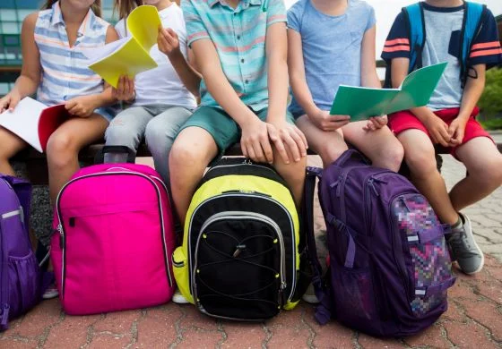 Photograph of a group of children sitting on a bench, holding their backpacks,