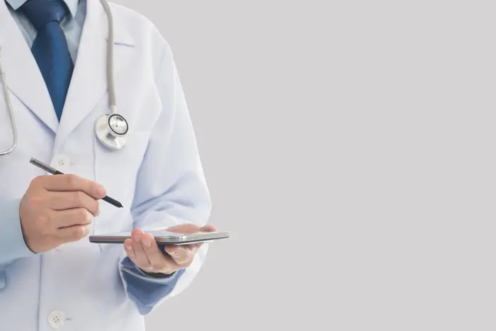 A doctor in a white lab coat and stethoscope is seen holding a tablet and writing on it.