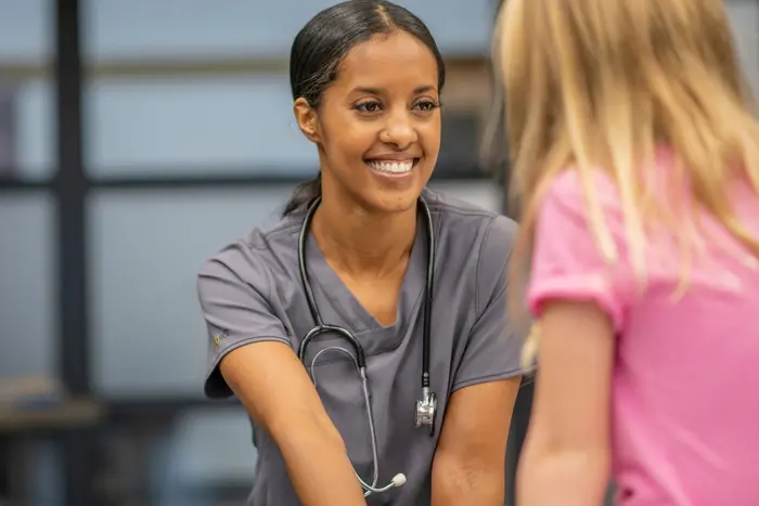 Photograph of a school nurse in grey scrubs and a stethoscope who is attentively conversing with a student in pink, providing care and support.