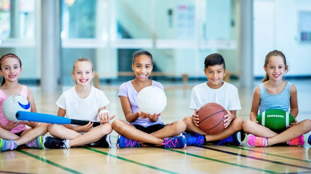 Image of five children smiling, each one is holding a different sporting item. One is holding a soccer ball, one is holding a blue foam bat, one is holding a volley ball, one is holding a basketball, and one is holding a green football.