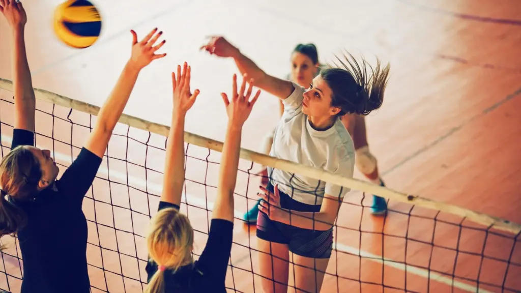 Photograph of four women playing volleyball. They are jumping over the net to hit the ball.