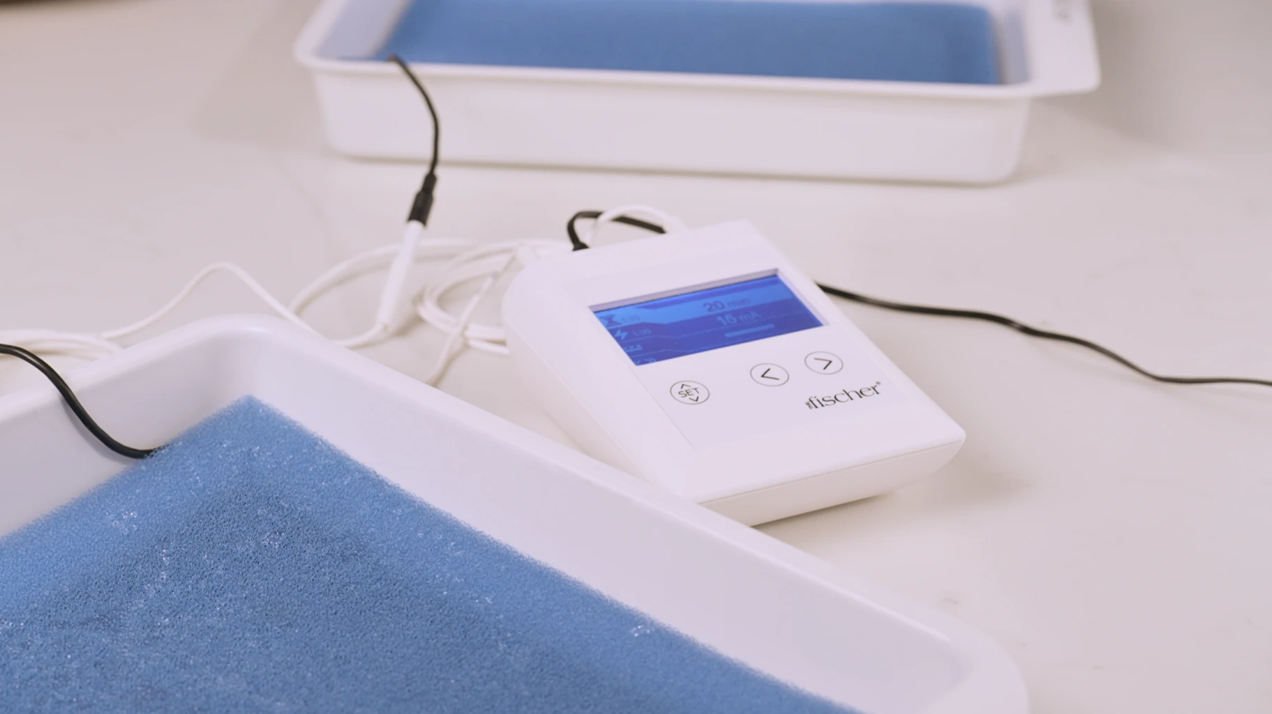 A photograph highlighting RA Fischer Co.'s original metal-free iontophoresis device, "The Fischer," specifically designed to treat hyperhidrosis, is displayed on a level surface. This device is seamlessly connected to two white water bath trays, each containing blue pH-balancing foam and featuring silicone-graphite electrodes, all neatly arranged atop a white table.