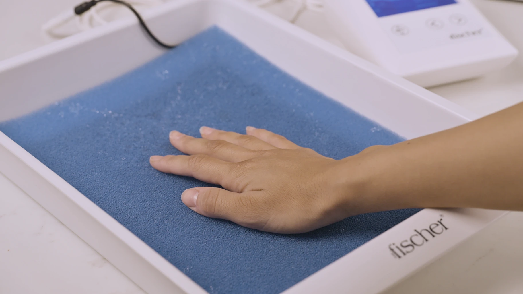 An individual effectively managing palmar (hands) hyperhidrosis utilizes RA Fischer Co.'s original metal-free iontophoresis device, "The Fischer." Their hand is comfortably placed within one of the white water bath trays containing blue pH-balancing foam and equipped with silicone-graphite electrodes, demonstrating a comprehensive solution for treating their condition.