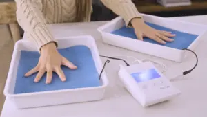 In a cozy home setting, a young woman sits contentedly,she addresses her palmar (hands) hyperhidrosis using RA Fischer Co.'s original metal-free iontophoresis device, "The Fischer." Her hands are immersed in the white water bath trays, complete with blue pH-balancing foam and silicone-graphite electrodes, representing a comprehensive and effective approach to managing her condition.