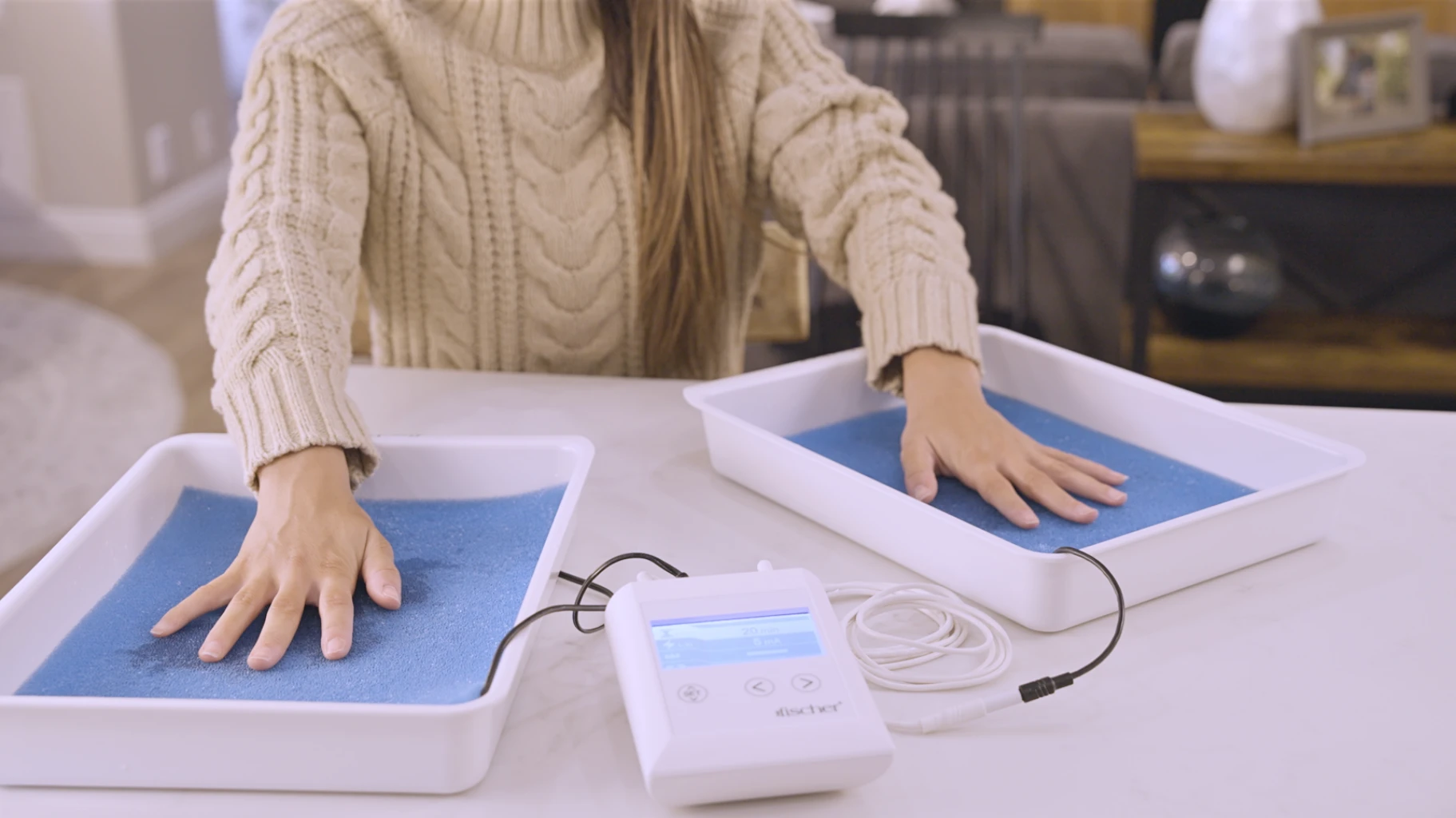 In a comfortable home environment, a young woman with brown hair sits with a warm smile while effectively managing her palmar (hands) hyperhidrosis (excessive sweat) using RA Fischer Co.'s original metal-free iontophoresis device, "The Fischer." Her hands are comfortably placed within the white water bath trays containing blue pH-balancing foam, along with silicone-graphite electrodes, providing a comprehensive and innovative solution for her condition.