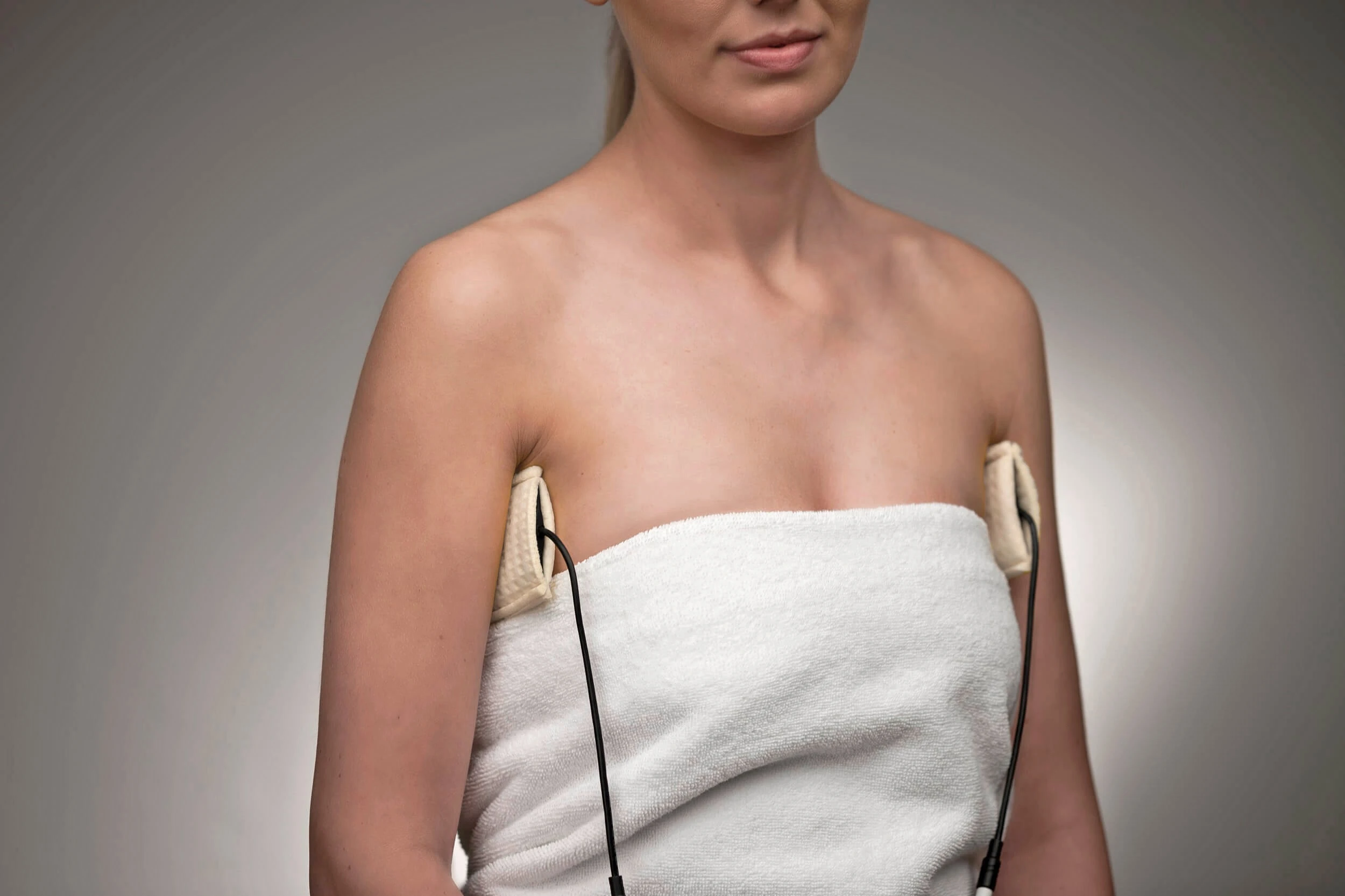 Photograph of a woman facing the camera wearing a white towel around herself. She has The Fischer's underarm attachments in her armpits and is using The Fischer to treat her axillary hyperhidrosis.