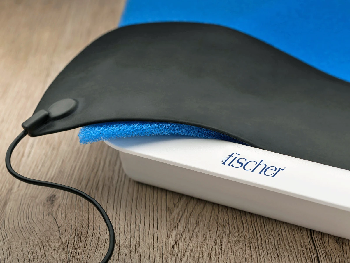 Close-up photograph of the white water bath trays and black, metal-free silicone electrodes for RA Fischer's "The Fischer" Device with ph-balancing foam inside the tray. The tray is sitting on a wooden table.