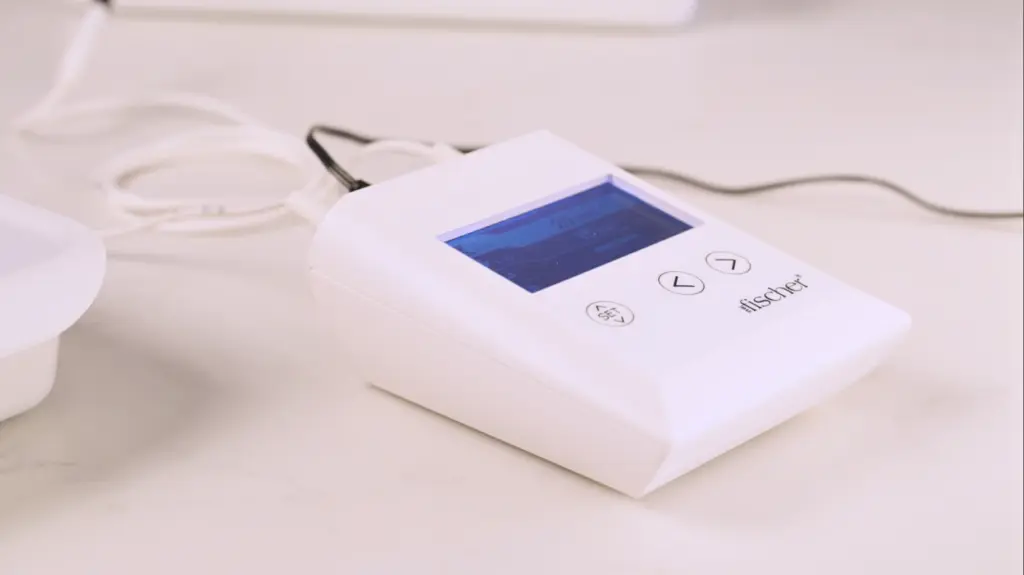 Image: A close-up photograph showcases the white, rectangular 'main control unit' of RA Fischer's 'The Fischer' Device, an advanced iontophoresis device meticulously engineered for effectively treating hyperhidrosis or excessive sweating. This device's distinguishing features include its white rectangular shape with a blue and grey screen. On the left side, there is a 'SET' button, and on the right side, two arrow buttons provide user control. The Fischer device's logo is discreetly located in the bottom right corner.