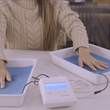A woman wearing a light brown sweater putting her hands into the Fischer iontophoresis device water bath trays to treat palmar hyperhidrosis aka excessive sweating of the hands