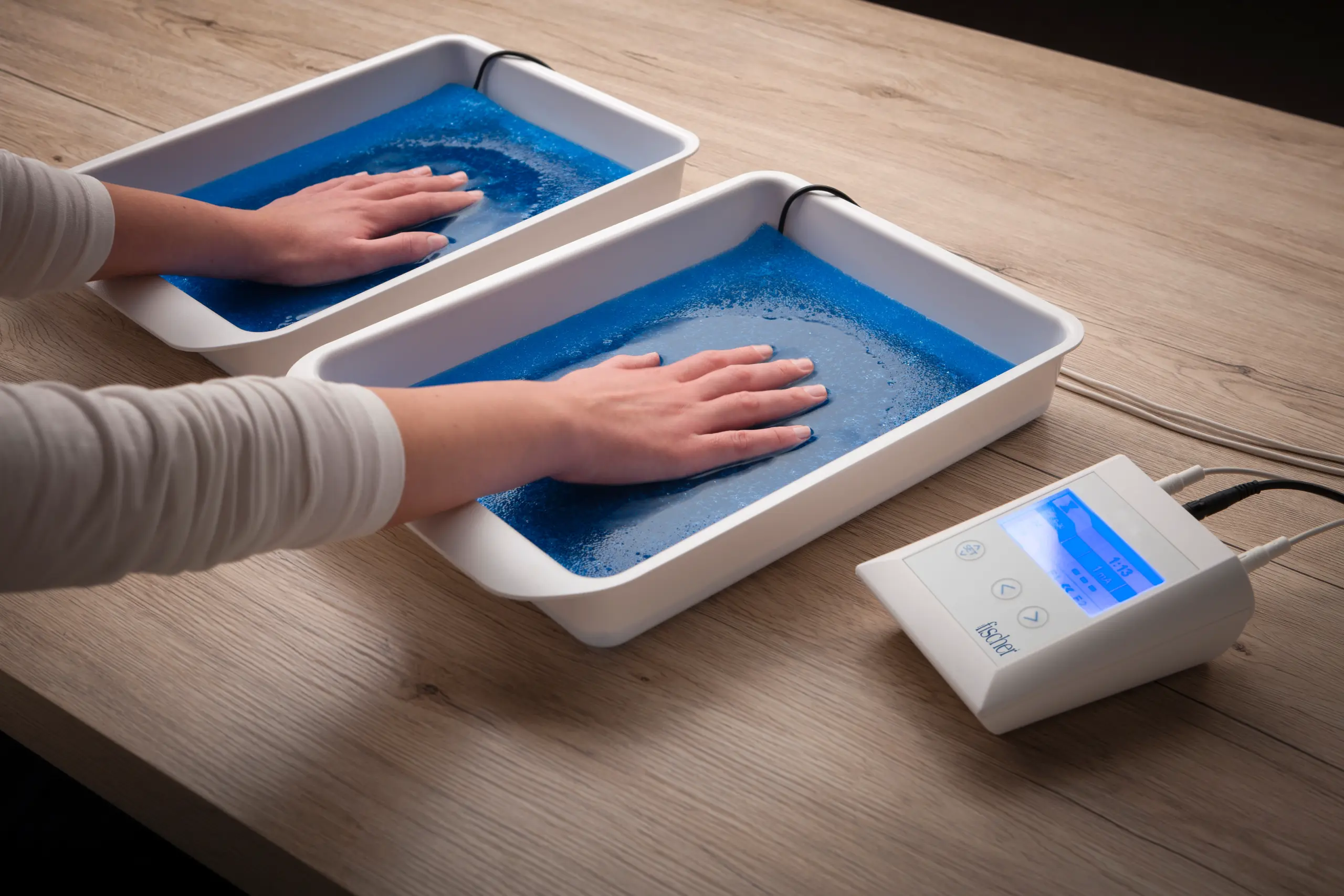 In this overhead photograph, we exclusively focus on the arms and hands of an individual facing forward while effectively addressing palmar hyperhidrosis, characterized by excessive hand sweating. They are utilizing RA Fischer Co.'s The Fischer iontophoresis device, strategically positioned on a white table. The scene highlights the device's key elements: white water bath trays, black metal-free silicone electrodes containing pH-balancing foam, all set against a plain white background. The Fischer iontophoresis device features a white rectangular design with a blue and grey screen, including buttons labeled "SET" and directional arrows, while the Fischer logo is discreetly located in the bottom right corner.