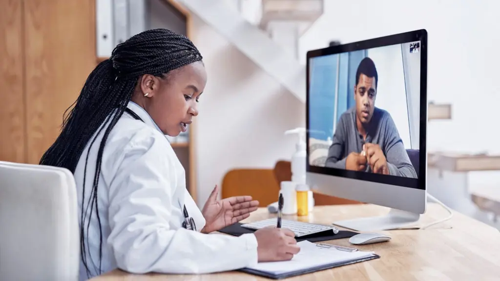 Picture of a telemedicine appointment from RA Fischer, a doctor talks to a patient virtually. Patient is pictured on the computer screen. Doctor is wearing a white lab coat and is writing on a clipboard.