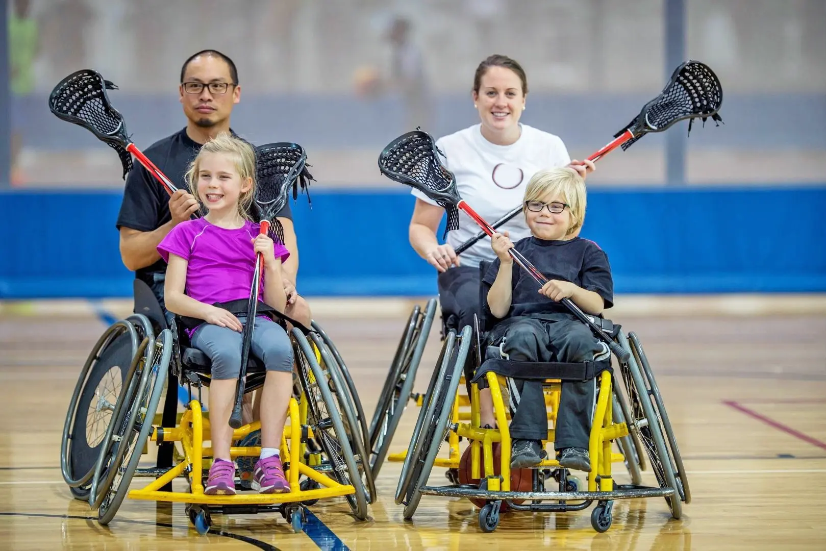 Picture of two younger patients and their parents participating in wheelchair lacrosse, in yellow wheelchairs, holding lacrosse sticks