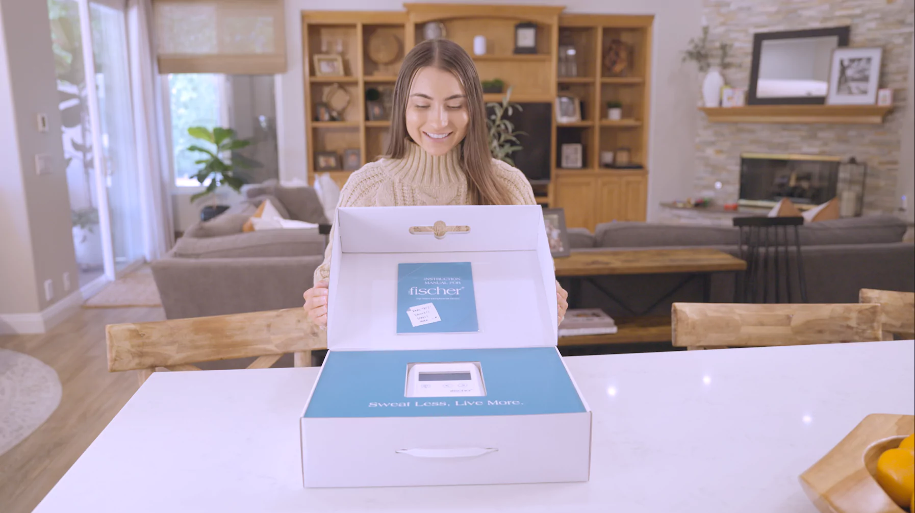 A young woman with brown hair standing at a table in a cozy-looking home. She is smiling and opening a box for RA Fischer Co.'s "The Fischer" iontophoresis device, made for treating hyperhidrosis (Excessive sweating).