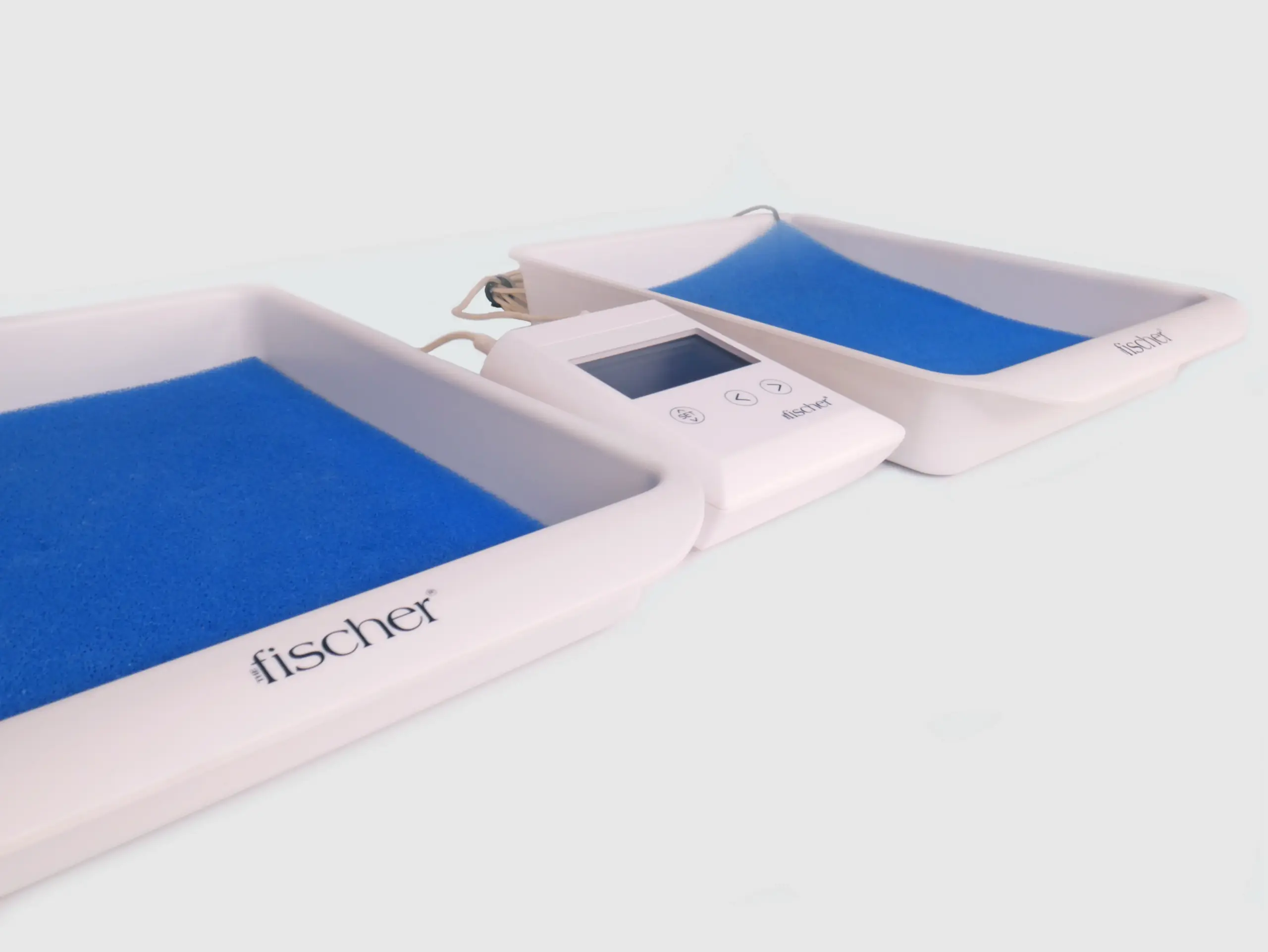 Angled photograph showcasing the white, rectangular 'main control unit' of RA Fischer's 'The Fischer' Device—an iontophoresis device designed for the treatment of hyperhidrosis (excessive sweating). The Fischer iontophoresis device is characterized by its white and rectangular shape with a blue and grey screen. On the left side of the device, there is a button labeled 'SET,' while two buttons on the right are represented by arrows. The Fischer device logo is placed in the bottom right corner. It is in between the water bath trays that have the blue ph-balancing foam and metal-free electrodes inside. There are cords visible behind the device. Iontophoresis Device for Sweating