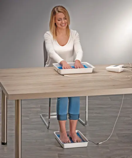 Image of a woman sitting at a table and using the Fischer iontophoresis device to treat hyperhidrosis. Two hands are in one tray and her feet are in the other. The main control unit sits on the table to her left