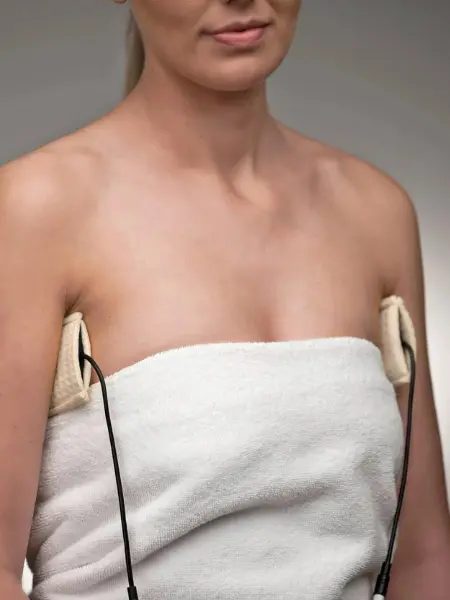 Image of a woman wrapped in a white towel. She is using the underarm attachments (UAs) for The Fischer iontophoresis device to treat hyperhidrosis
