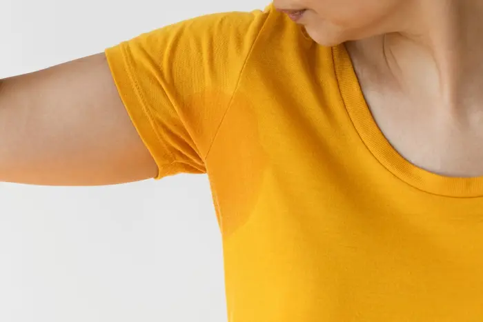 A woman in a yellow shirt holds her arm, examining sweat stains on her clothing which could be caused by hyperhidrosis (axillary hyperhidrosis).