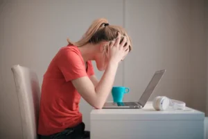 Photograph of a frustrated girl in a red shirt sitting at a desk, her head in her hands, while looking at a laptop.