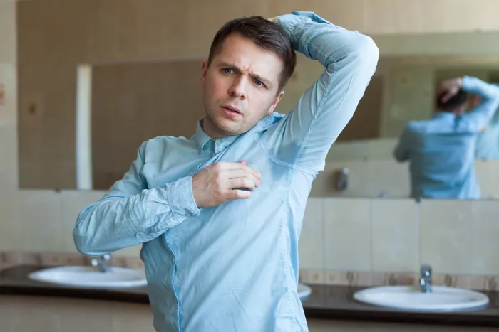 In front of a bathroom mirror, a man wearing a blue shirt scrutinizes a sweat stain on his armpit, which could be from axillary hyperhidrosis